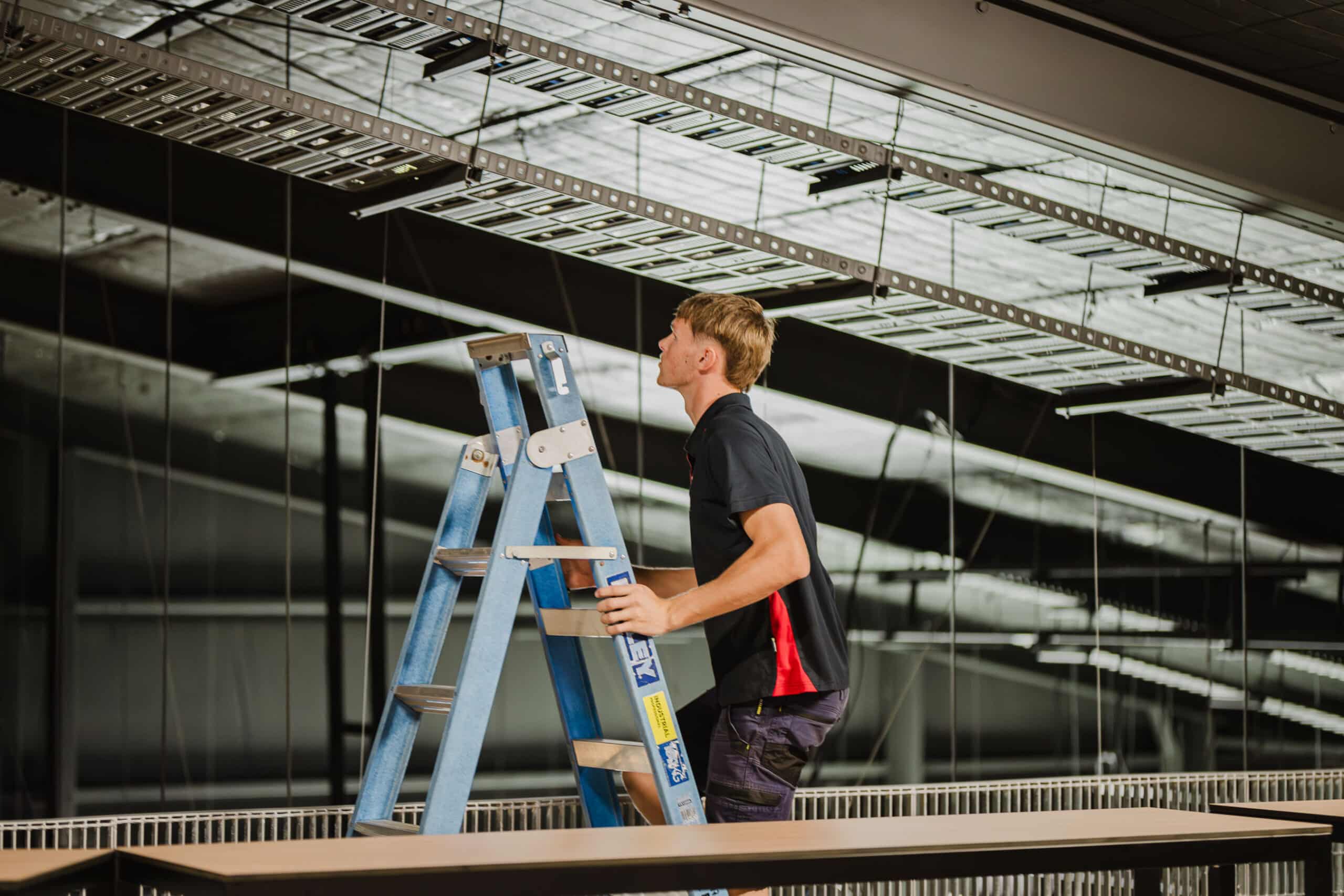 Electrician on a ladder inspecting overhead cable trays during a commercial electrical installation.